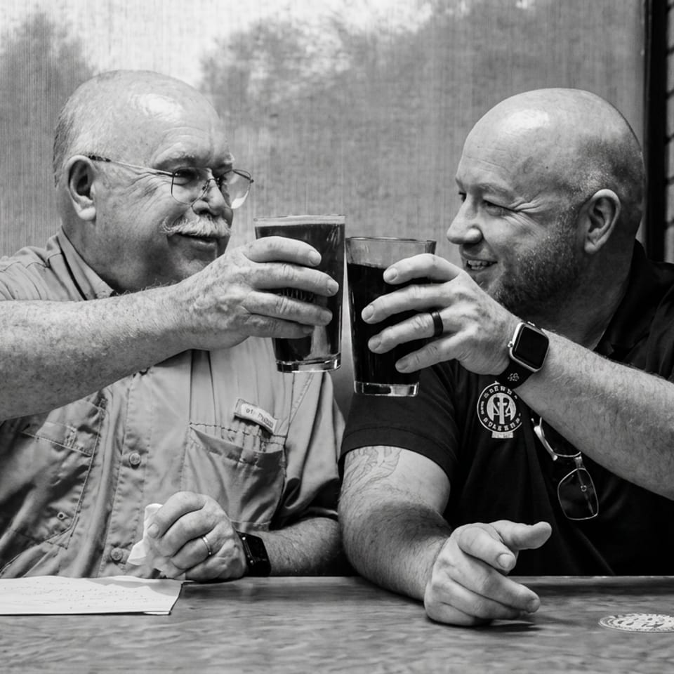 Two men smile and toast with dark beers at a restaurant table beside a brick wall.