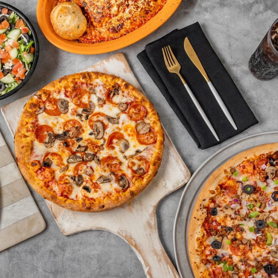 Overhead spread of pepperoni and mushroom pizzas, a loaded vegetable pizza, salad, baked pasta, utensils, and iced soda.