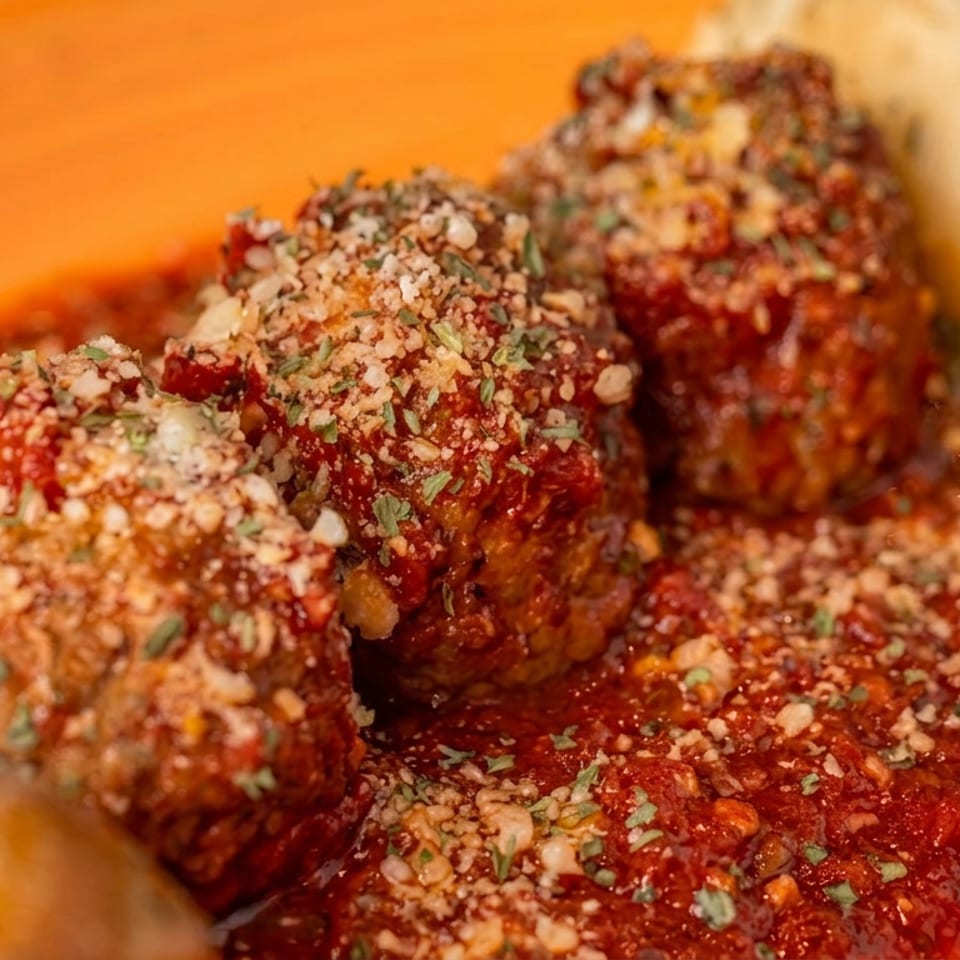 Close-up of meatballs in tomato sauce, topped with grated cheese and herbs, served on an orange plate with toasted bread.